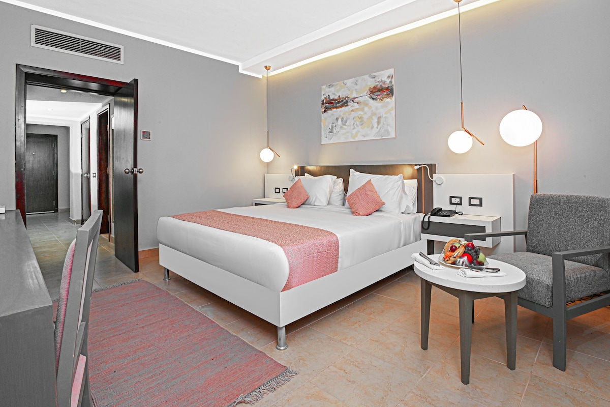 A modern hotel room with a large bed, a desk, and a chair. There is a painting on the wall and a small table with fruit on it.