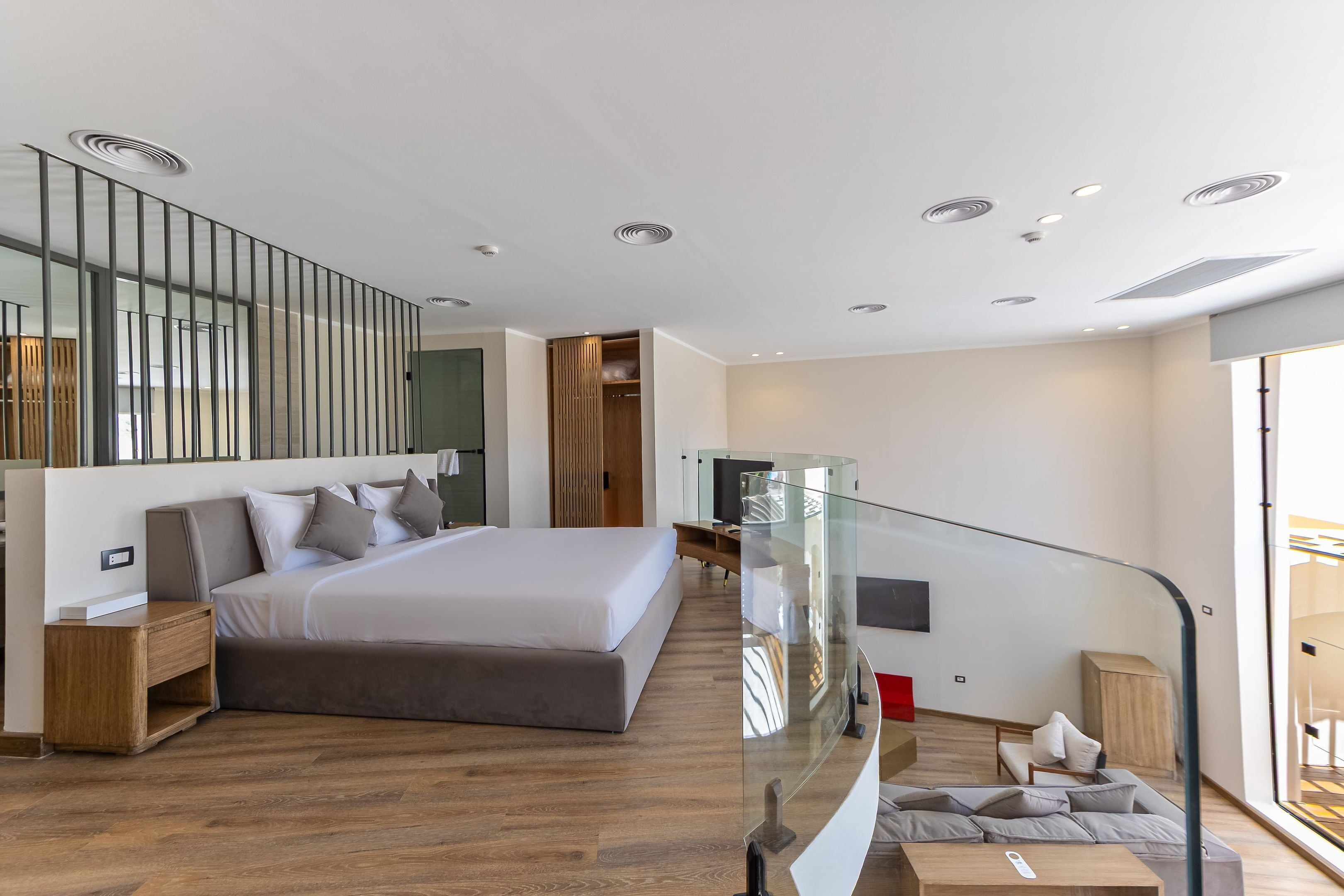 A modern bedroom with a large bed, wooden flooring, a glass railing, and a view of the outdoors.