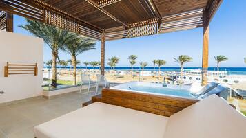 A room with a wooden ceiling, a bed, a view of the beach, and a pool area.
