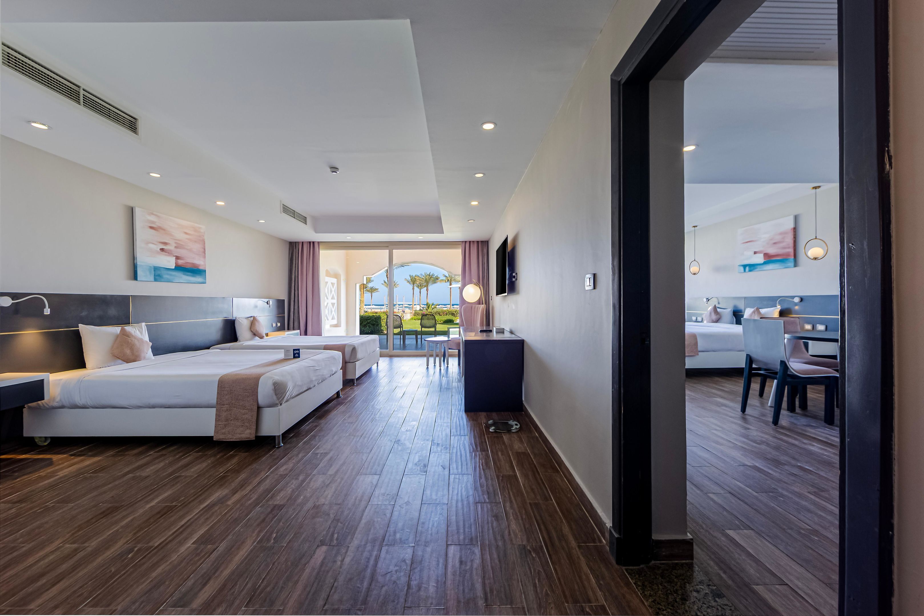 A modern hotel room with two beds, a dining area, and a view of the outdoors.