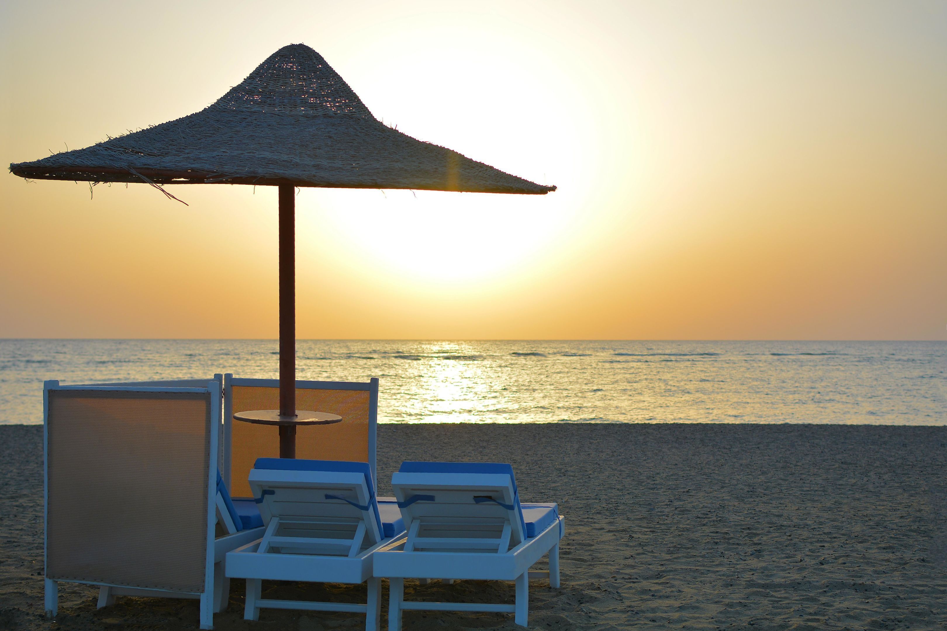A beach scene with a thatched umbrella, two lounge chairs, and a view of the ocean at sunset.