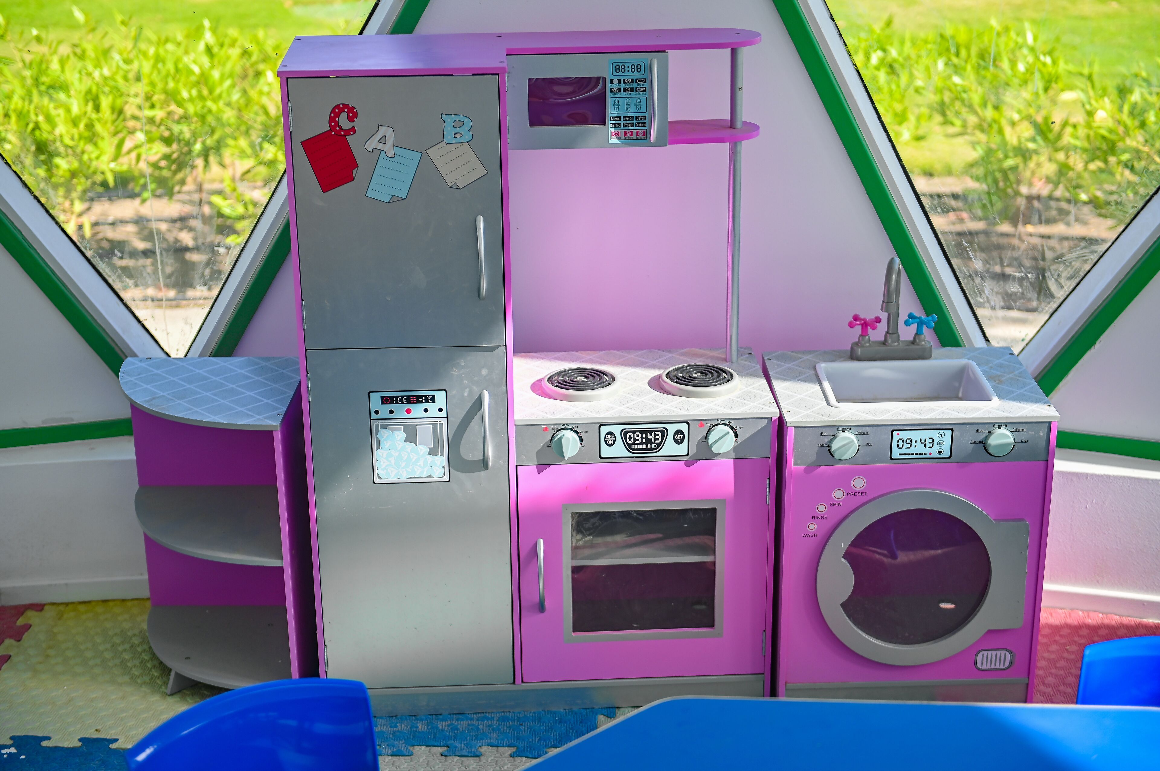 A children's play kitchen with a refrigerator, oven, and sink, set against a window with a view of a garden.