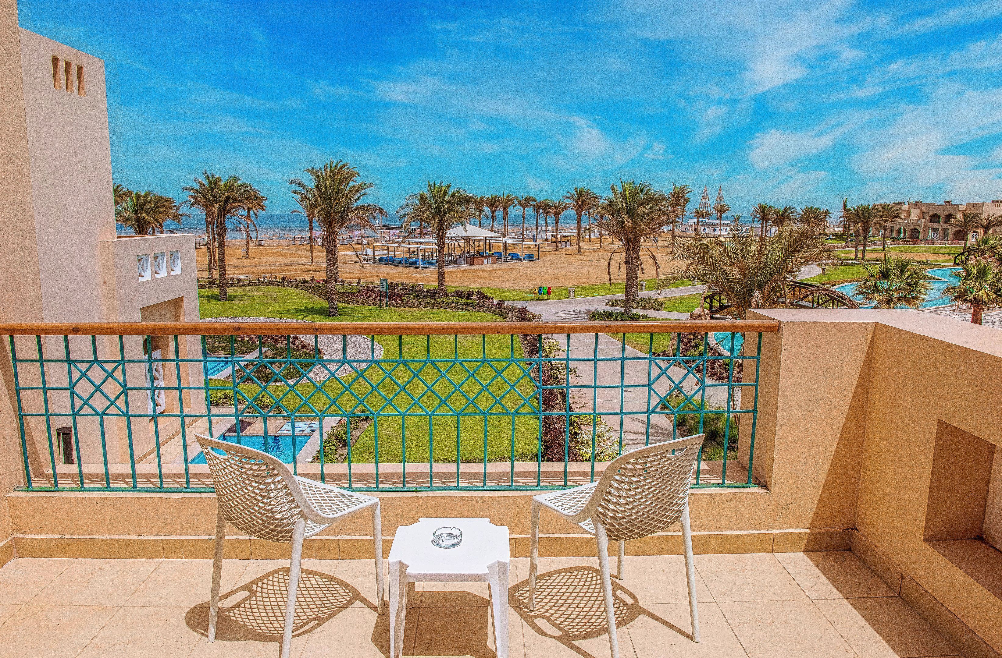 A balcony with two white chairs and a small table, overlooking a pool area with palm trees and a clear blue sky.