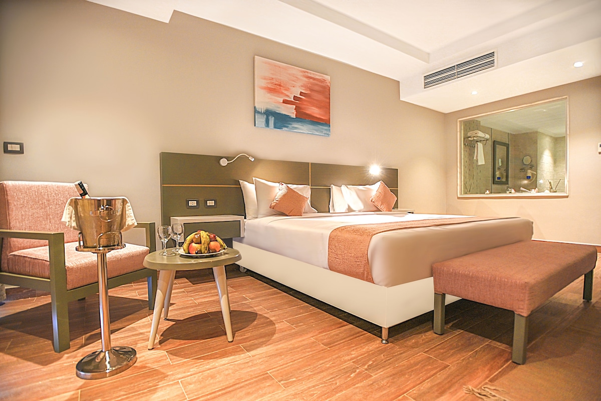 A hotel room with a bed, a bedside table with a fruit bowl, a chair, a bench, and a large mirror.