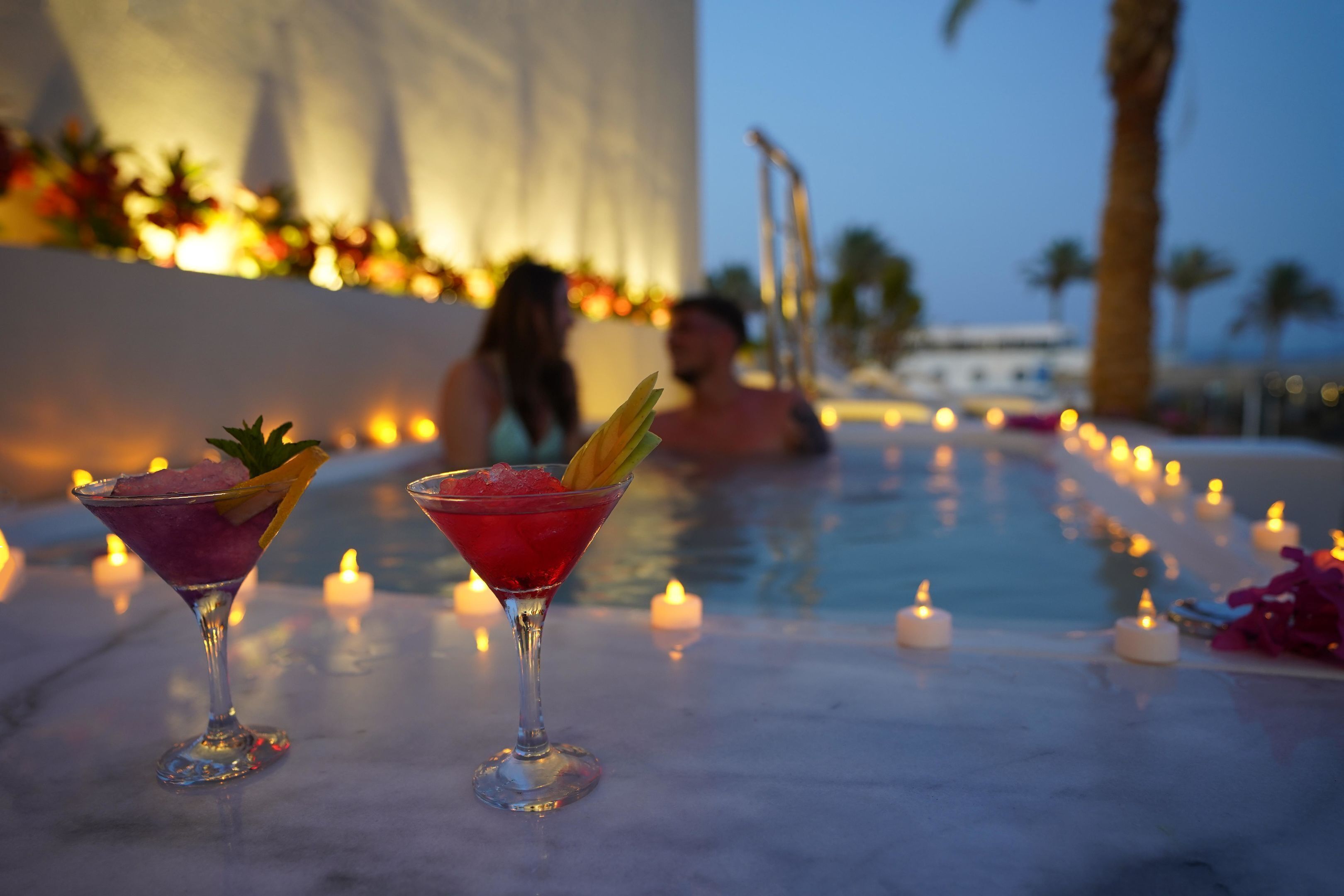 Two cocktails on a marble table with candles around a pool area.