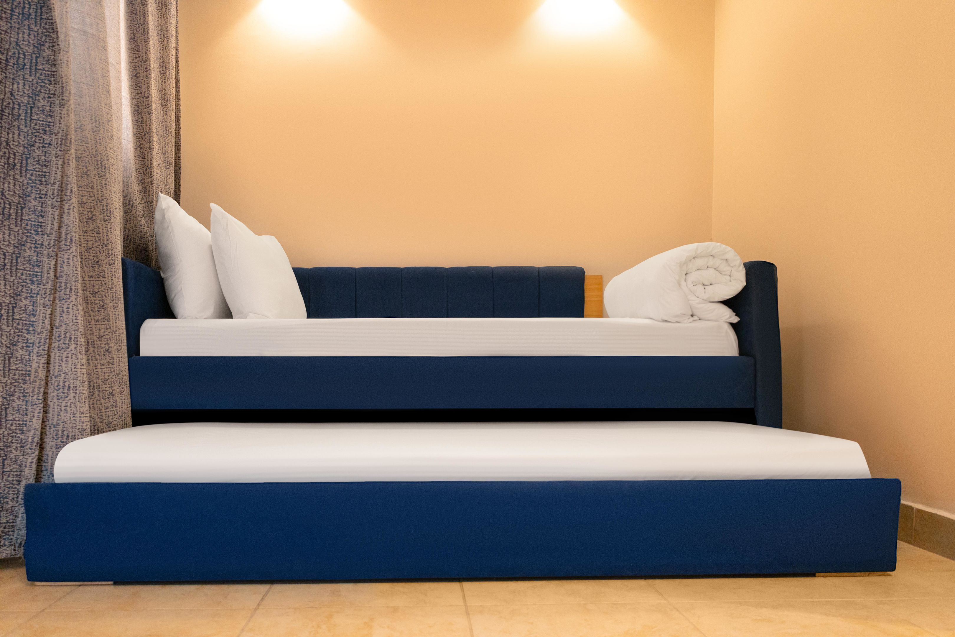 A double bed with a blue headboard and white bedding, a rolled towel, and a curtain on the left.