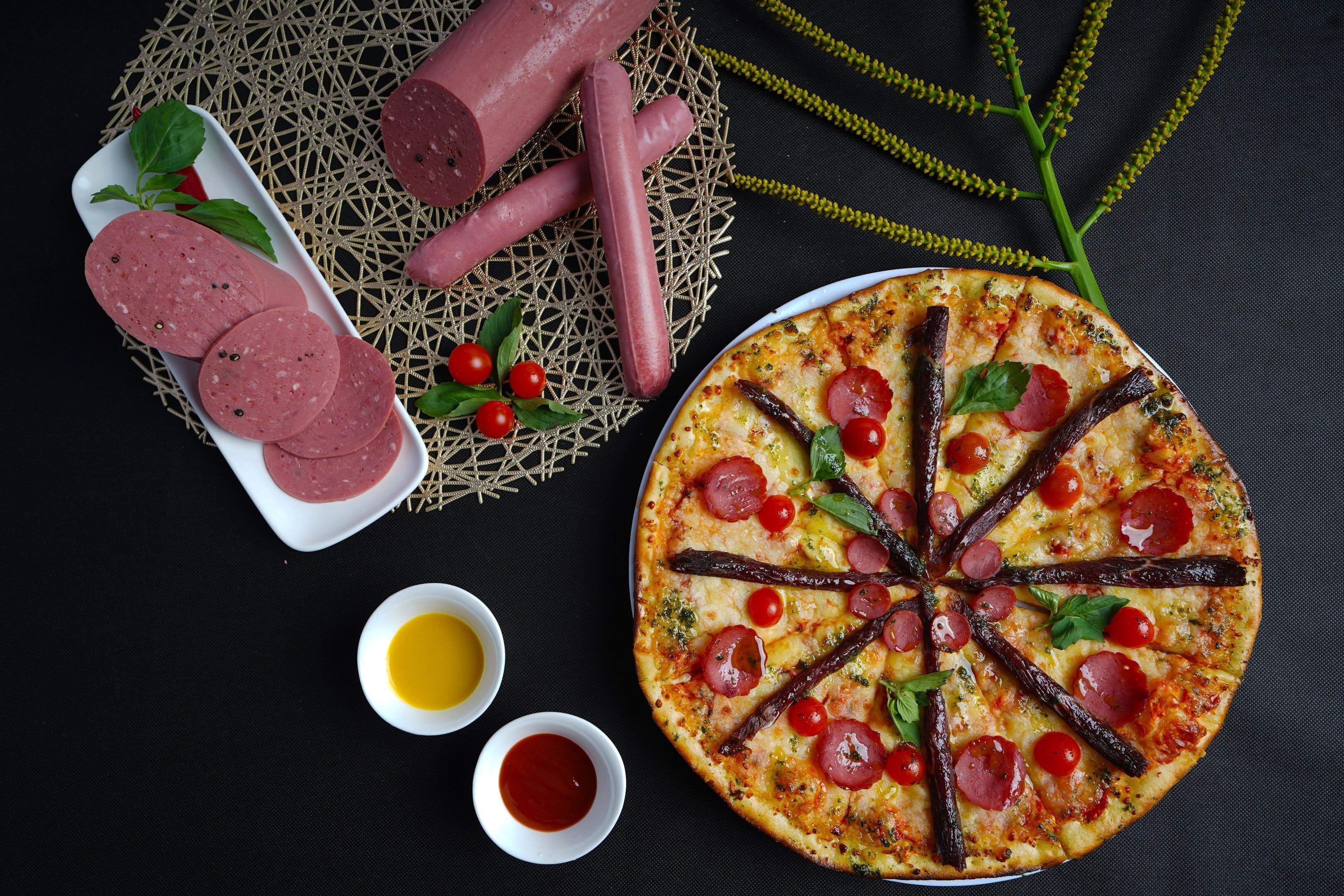 A pizza with pepperoni and sun-dried tomatoes, accompanied by sliced sausages and two small bowls of sauce.