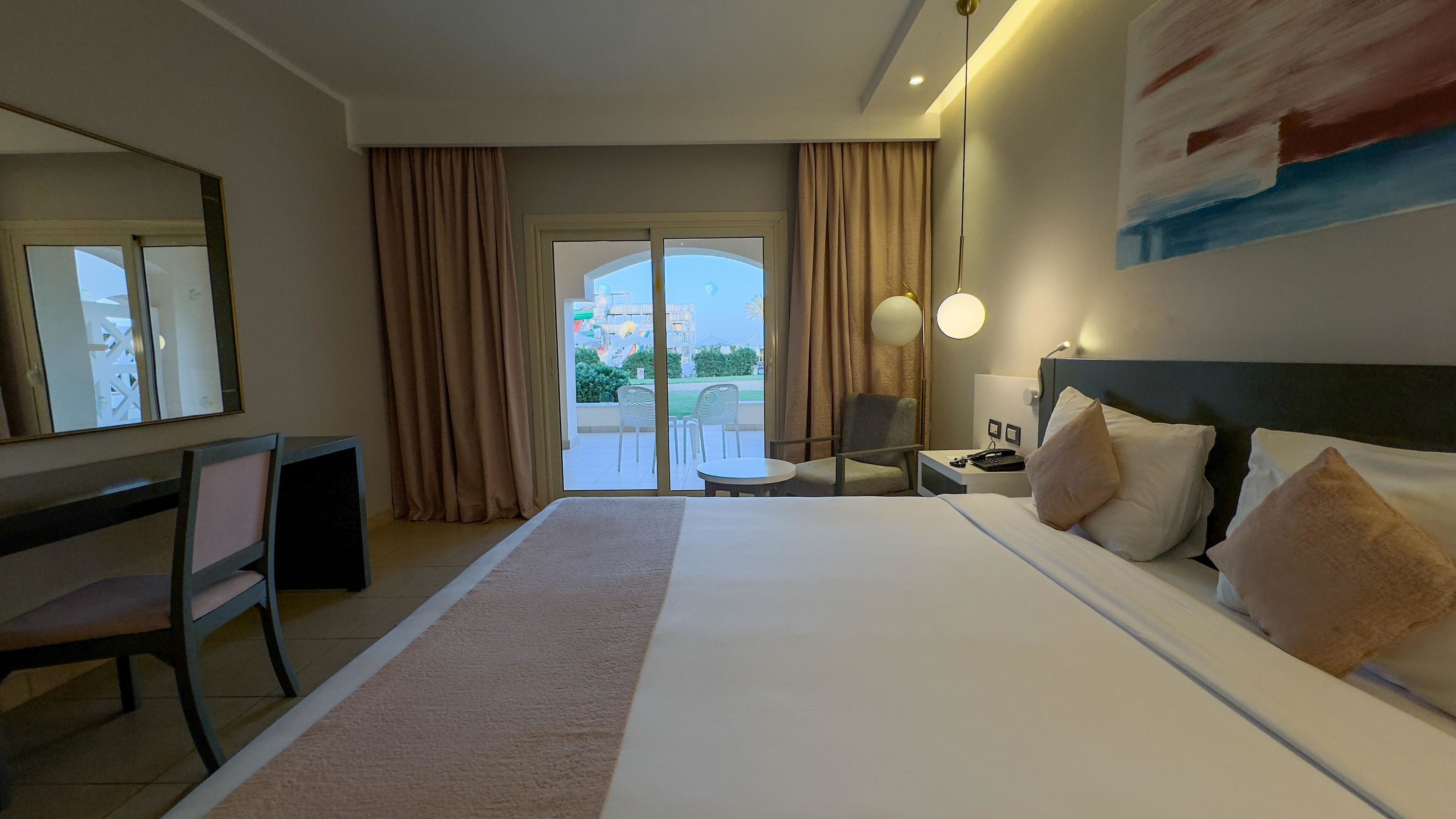 A hotel room with a large bed, a desk, a chair, a nightstand, and a view of the outdoors through a glass door.