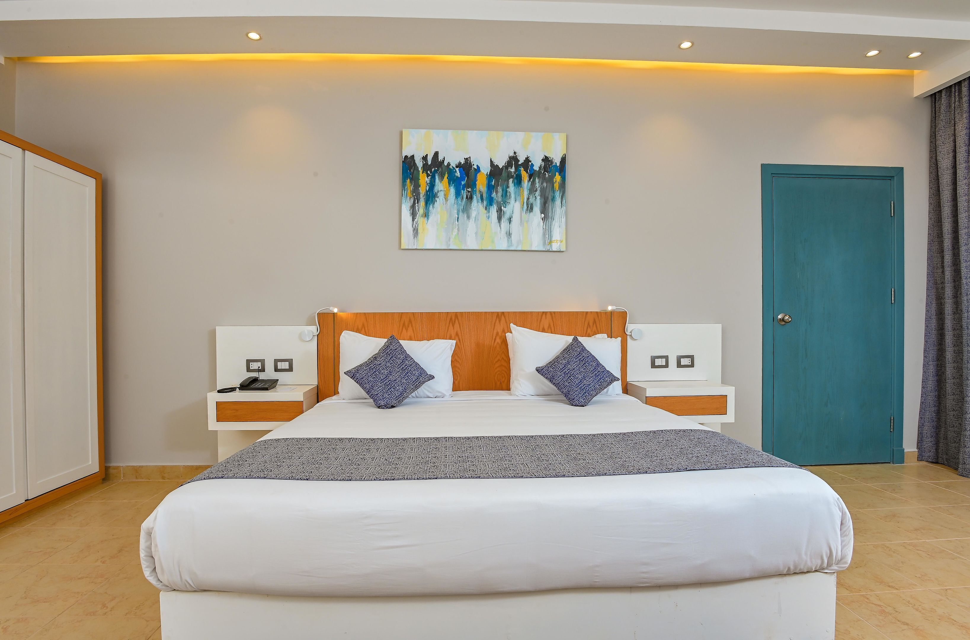 Una habitación de hotel moderna con una cama grande, un cabecero de madera, dos mesitas de noche con lámparas, un cuadro en la pared y una puerta color turquesa.