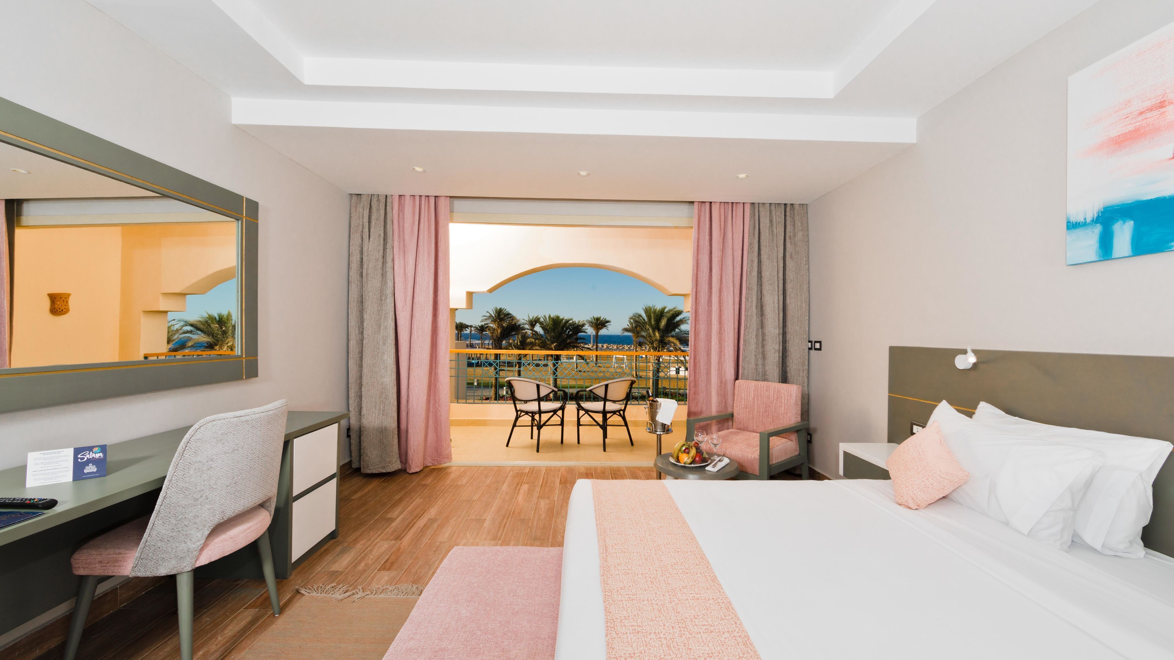 A hotel room with a bed, desk, chair, and a balcony with a view of palm trees and the sea.
