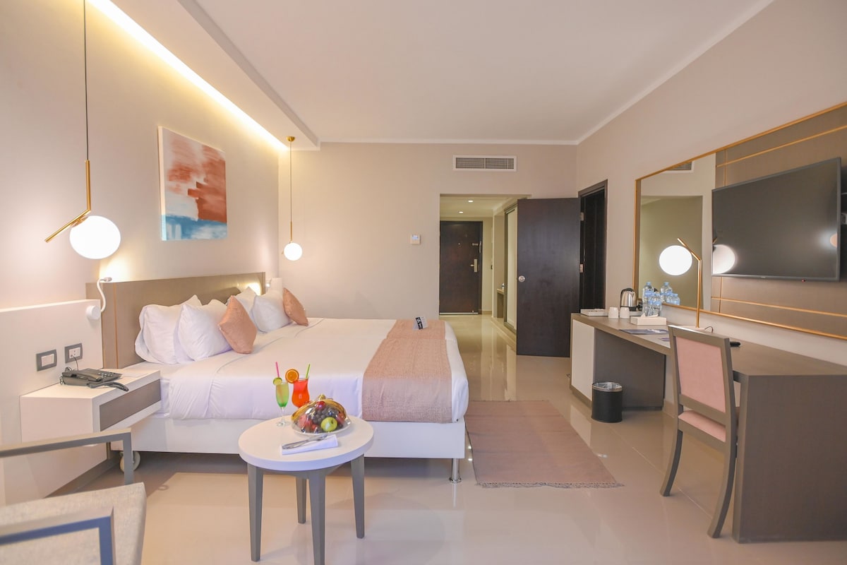 A modern hotel room with a neatly made bed, a bedside table with a phone and charger, a small round table with fruit and a drink, a desk with a chair, and a large mirror.