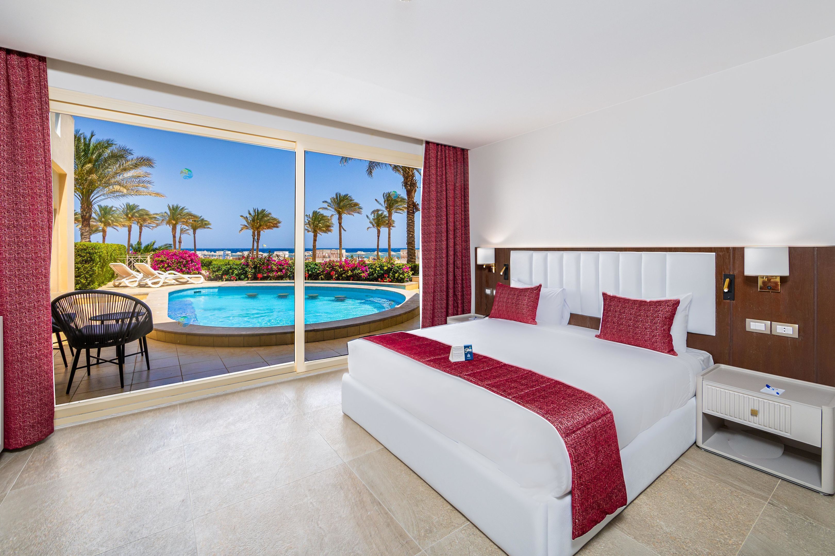 A hotel room with a large bed, a bedside table, a view of a pool and palm trees.