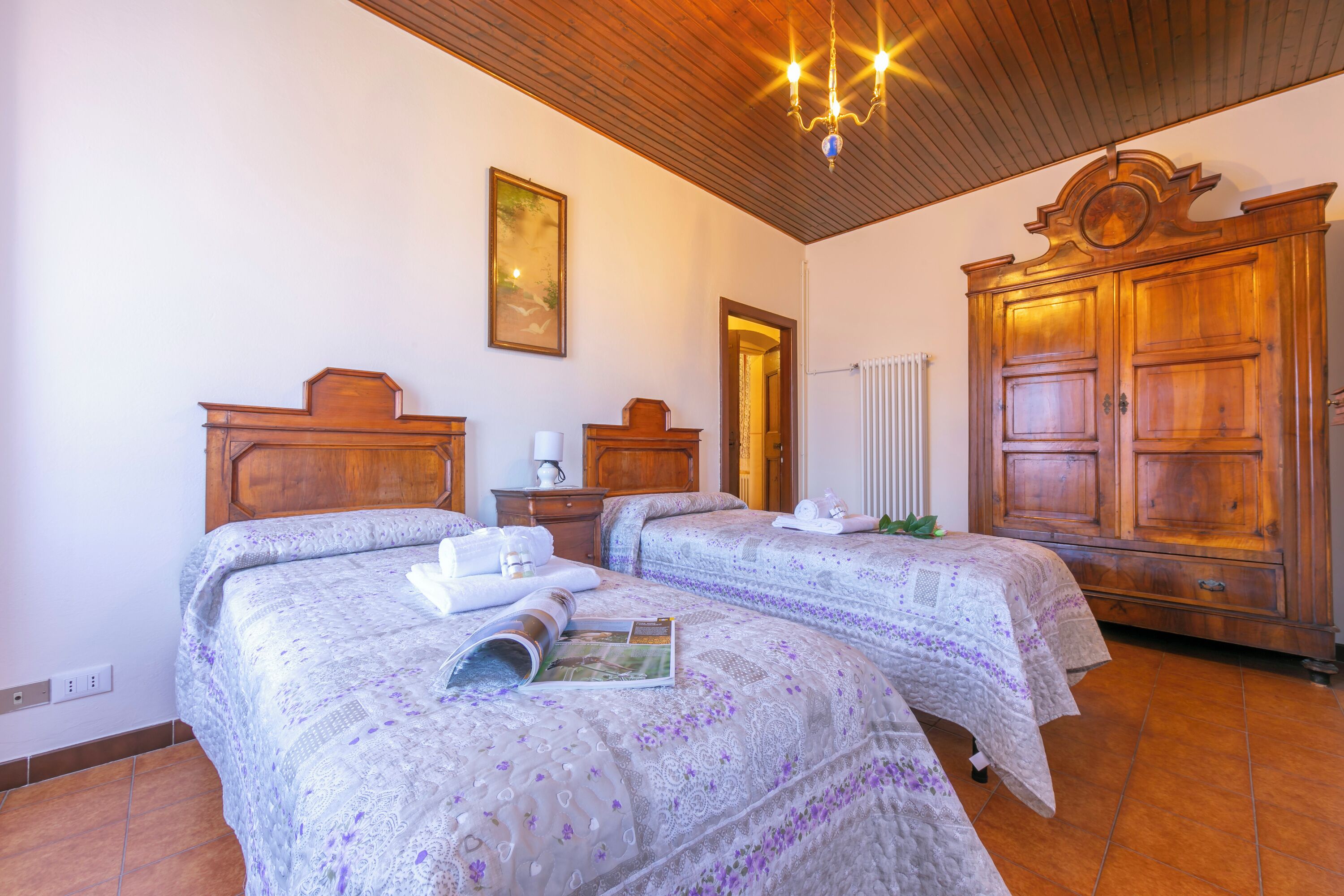 A room with two beds, wooden headboards, a chandelier, a radiator, and a large wooden wardrobe.