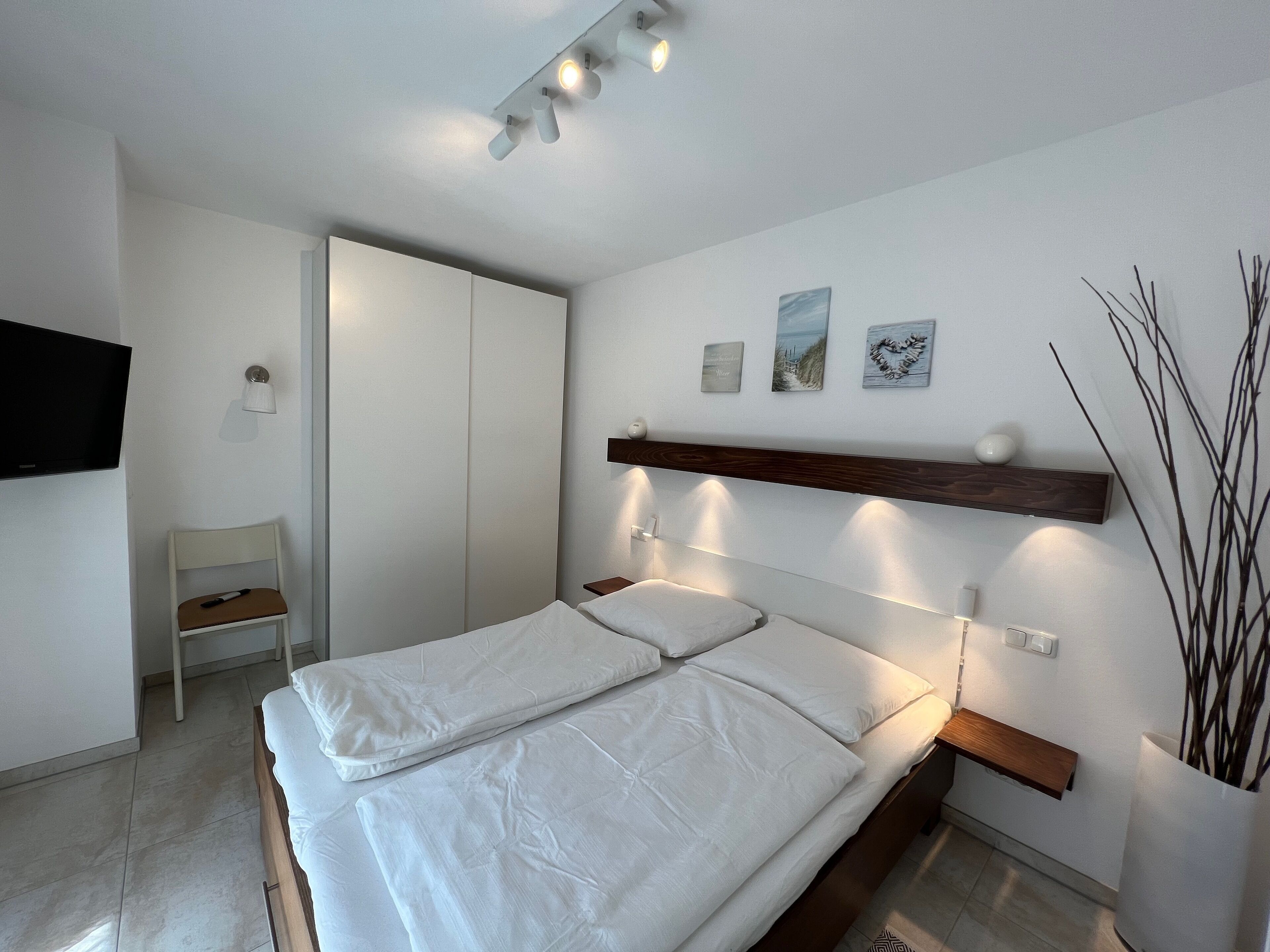Une chambre à coucher moderne, avec un lit, des tables de chevet, un téléviseur, une chaise et des œuvres d’art accrochées au mur.