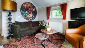 Un salon comprenant un canapé, une table basse ronde, une lampe, un tableau, un téléviseur et une fenêtre avec des rideaux.