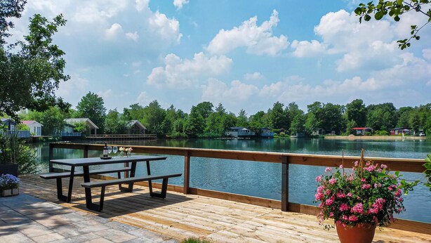 Une terrasse en bois avec des tables de pique-nique et un pot de fleurs, donnant sur un lac paisible, avec des maisons en arrière-plan.