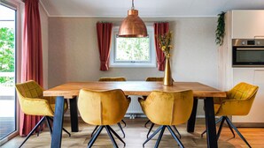 Un coin repas avec une table en bois, six chaises jaunes, un luminaire suspendu en cuivre, une fenêtre avec des rideaux et un espace cuisine équipé d’appareils électroménagers.