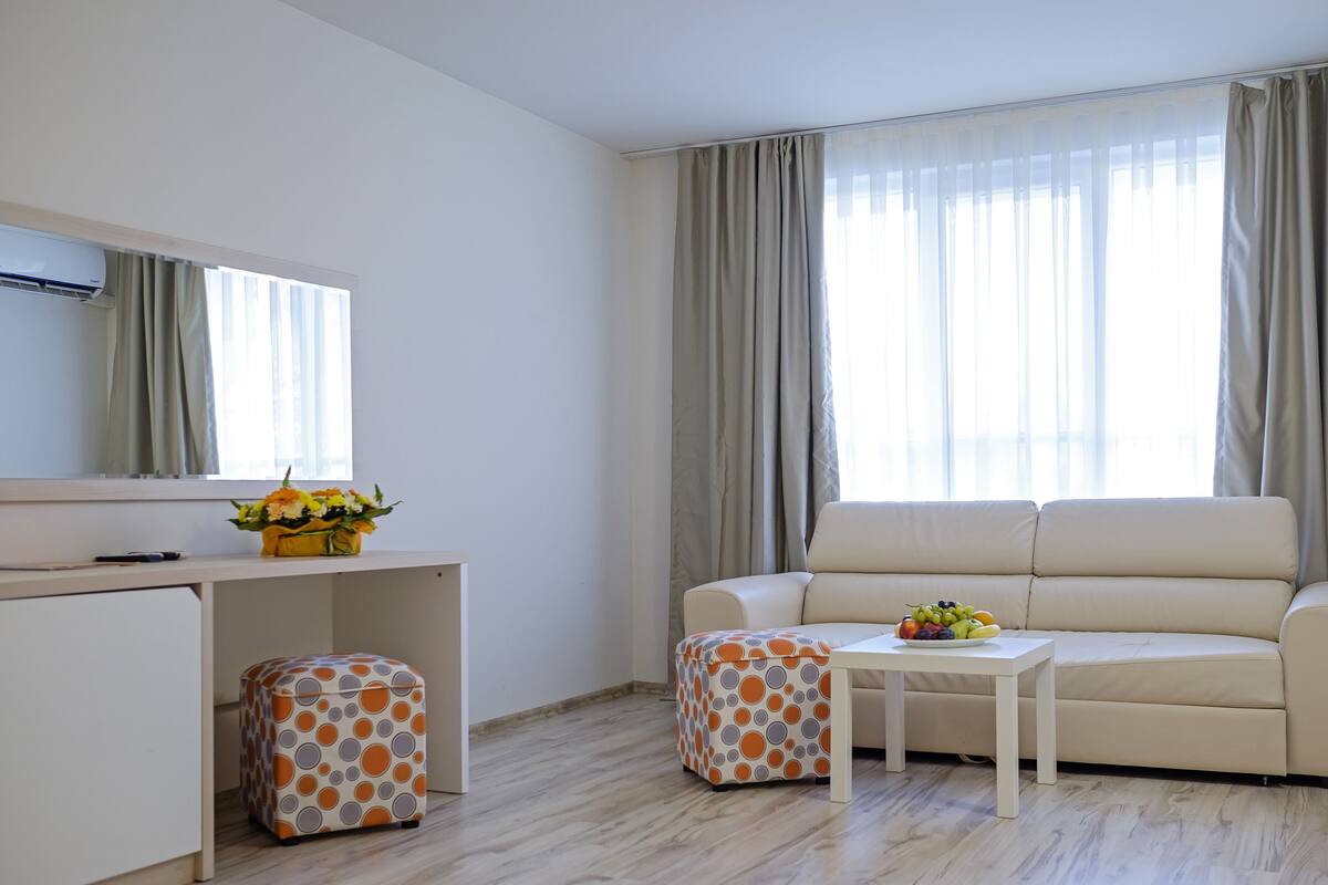 A modern living room with a sofa, a small table with fruit, a desk with a computer and flowers, and a patterned ottoman.