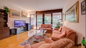A spacious living room with a large sofa, a coffee table, and a television. There are framed pictures on the walls and a plant on the left side. - Sweet Home (Lugano)