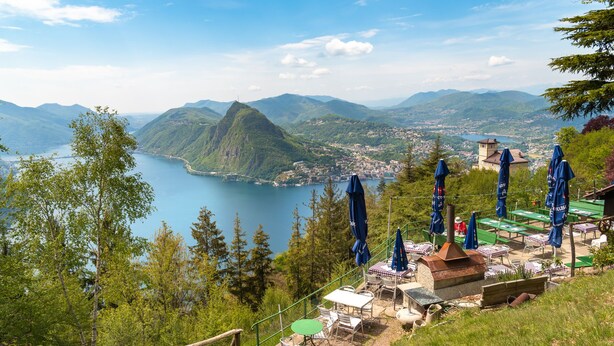 A scenic outdoor seating area with tables and chairs, overlooking a lake and mountains. - Sweet Home (Lugano)