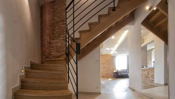 Una escalera con escalones de madera y barandillas negras que conduce a una habitación con pared de ladrillo y un espejo grande.