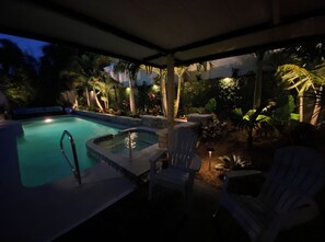 A hot tub with a pool, surrounded by chairs and palm trees under a covered area.