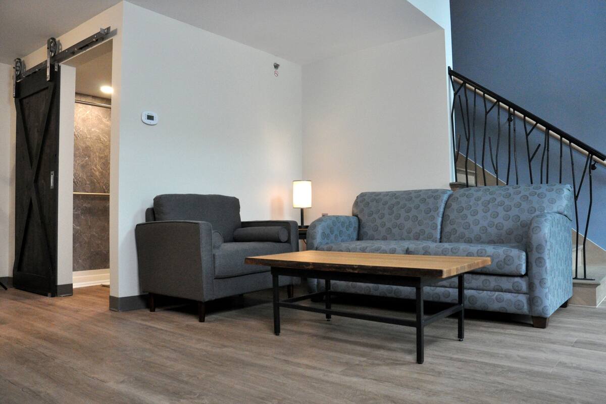 A living room with a grey sofa, a smaller armchair, a wooden coffee table, and a lamp. There is a sliding door leading to another room and a staircase with a metal railing.