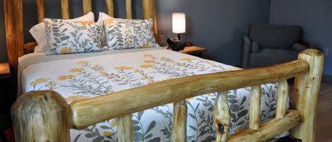 Una cama con estructura de madera, ropa de cama con motivos florales, una lámpara de noche y un sillón gris.