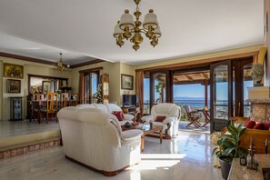 A spacious living room with a large sofa, armchairs, and a coffee table. A television is on a wooden stand, and there is a balcony with a view of the sea.