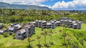 A modern resort with multiple buildings featuring dark stone exteriors and flat roofs, surrounded by lush greenery and a view of the ocean.