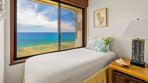 A bedroom with a large window offering a view of the ocean. There is a bed with a white bedspread and a pillow with a tropical leaf pattern. A wooden nightstand with a lamp and a decorative item is beside the bed.