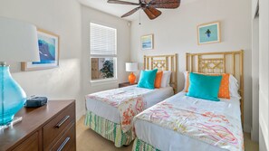 Une chambre avec deux lits, un ventilateur de plafond, une fenêtre avec des stores et un bureau avec une lampe.