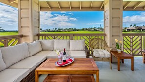 A covered outdoor seating area with a view of a golf course, a wicker chair, a wooden table with a tray of drinks, and a small table with flowers. - Pili Mai at Poipu #06E By Parrish Kauai- best Poipu location & AC - walk to shop (Koloa)
