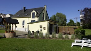 Una casa de dos pisos con techo inclinado, un porche delantero y un pequeño jardín con una parrilla y mobiliario exterior.