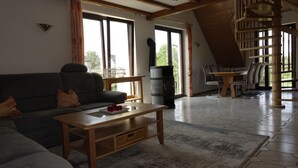 Una sala de estar con un sofá, una mesa de centro y una chimenea. Hay un comedor con una mesa y sillas. La habitación tiene ventanas grandes y una escalera de madera.