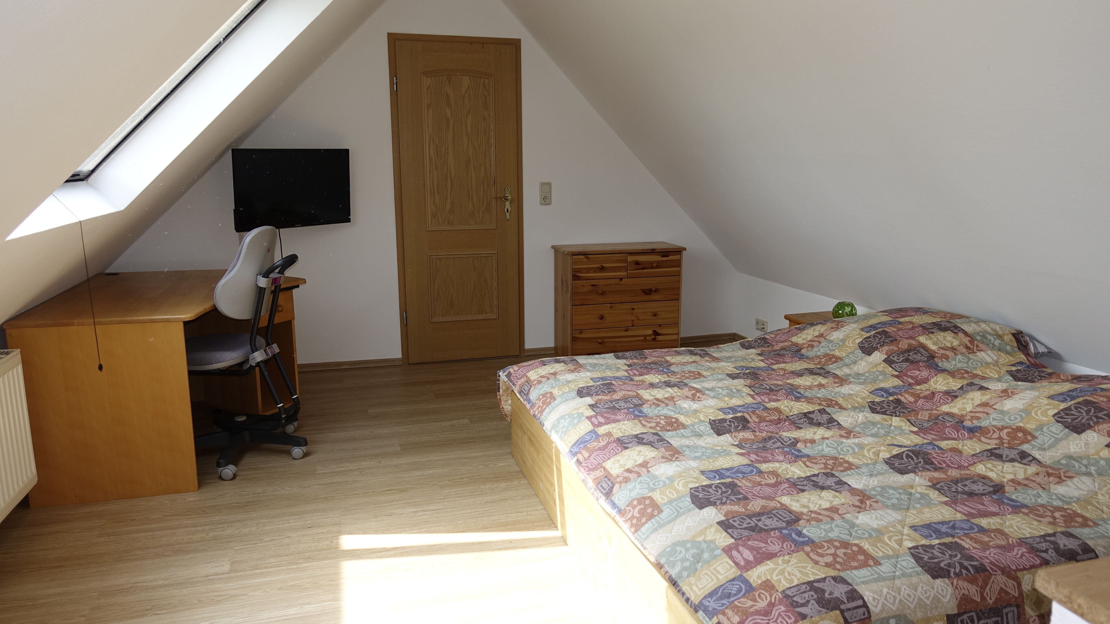 A bedroom with a sloped ceiling, a desk with a chair, a bed with a patterned quilt, a wooden wardrobe, and a television on the wall.
