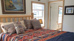 Un dormitorio con una cama de madera, almohadas estampadas, una colcha colorida, un cuadro sobre la cama y una ventana con persianas.