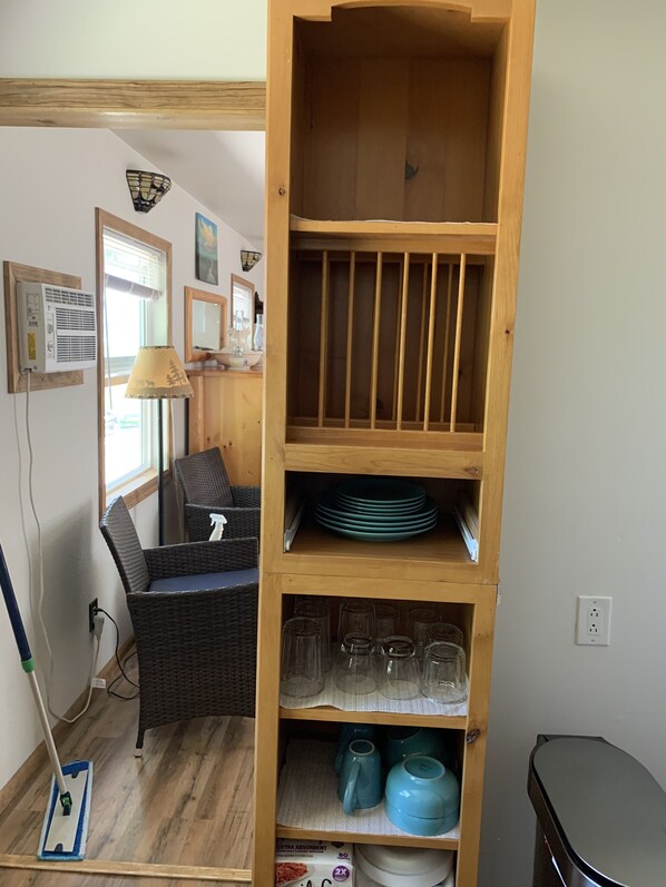 A room with a wooden shelving unit containing plates, glasses, and teapots. There is a wicker chair and a lamp near a window.