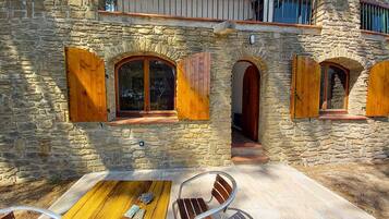 Un bâtiment en pierre doté de fenêtres en arc de cercle et de volets en bois, une terrasse avec une table et des chaises, et un balcon avec des balustrades.