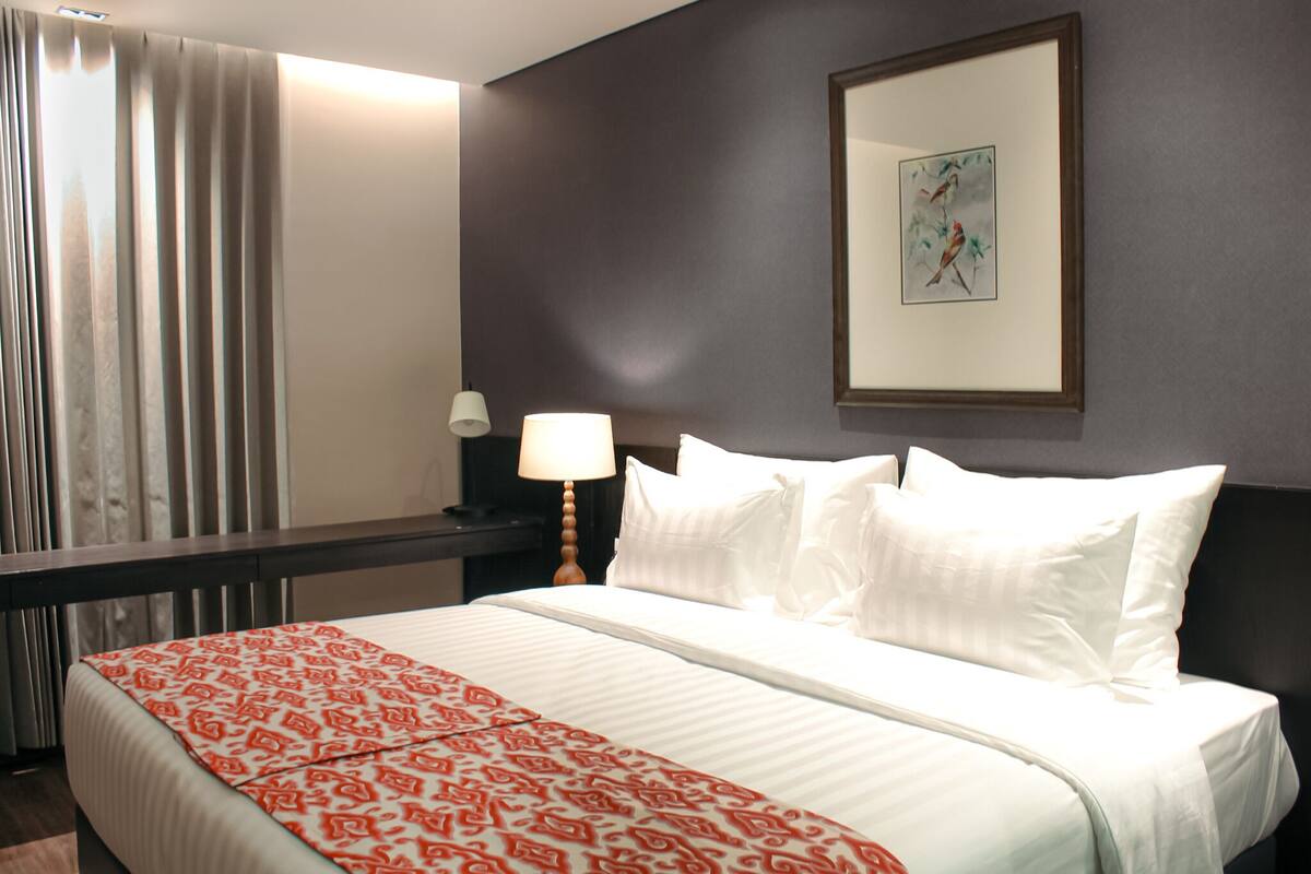 Une chambre d’hôtel avec un grand lit, une table de chevet avec une lampe, un tableau encadré au mur et une fenêtre avec des rideaux.