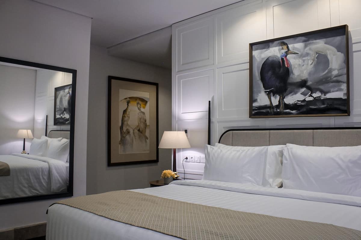 Une chambre d’hôtel avec un grand lit, une table de chevet avec une lampe, une œuvre d’art encadrée au mur et un tableau au-dessus du lit.