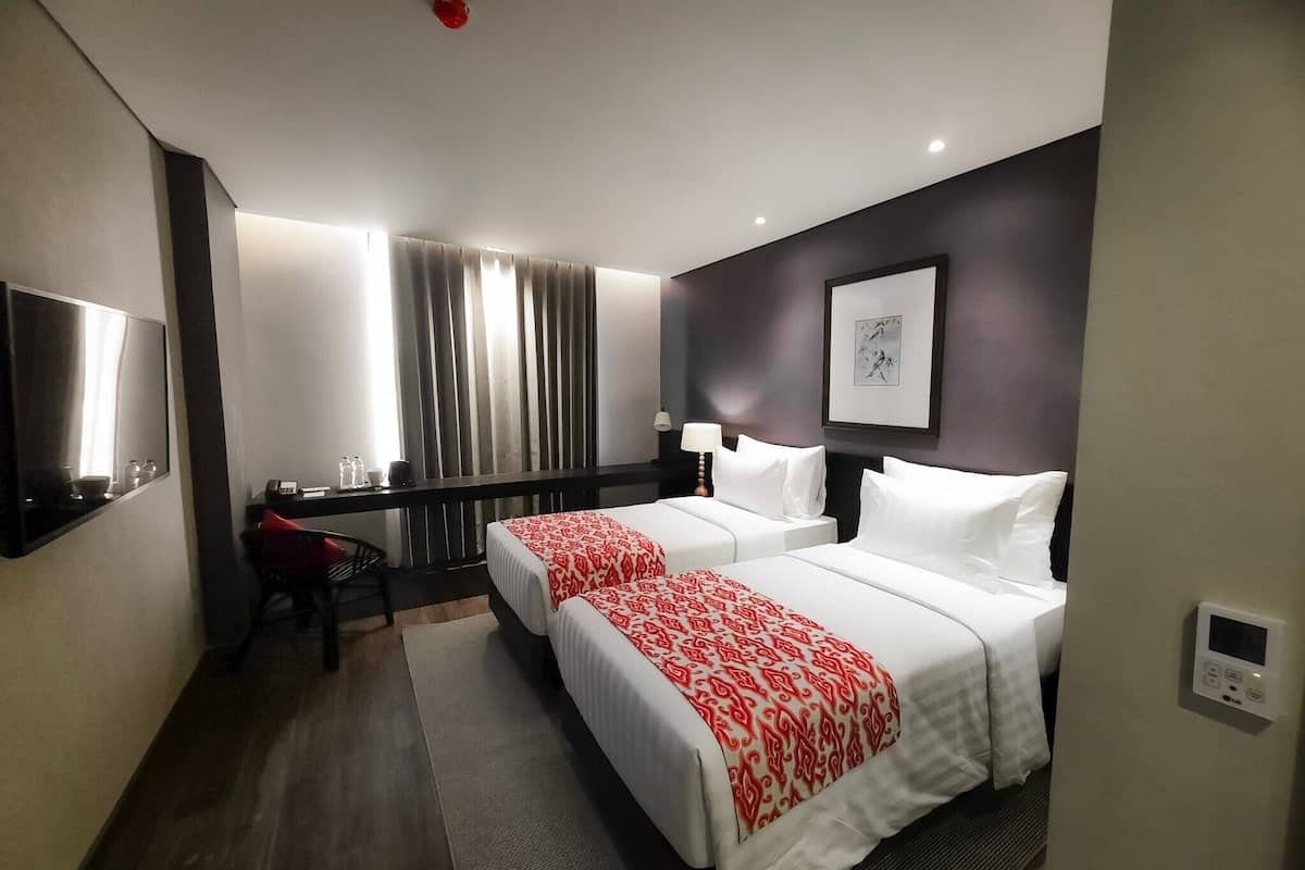 Une chambre d’hôtel avec deux lits, un bureau, une chaise, une télévision, une fenêtre avec des rideaux et un tableau encadré au mur.