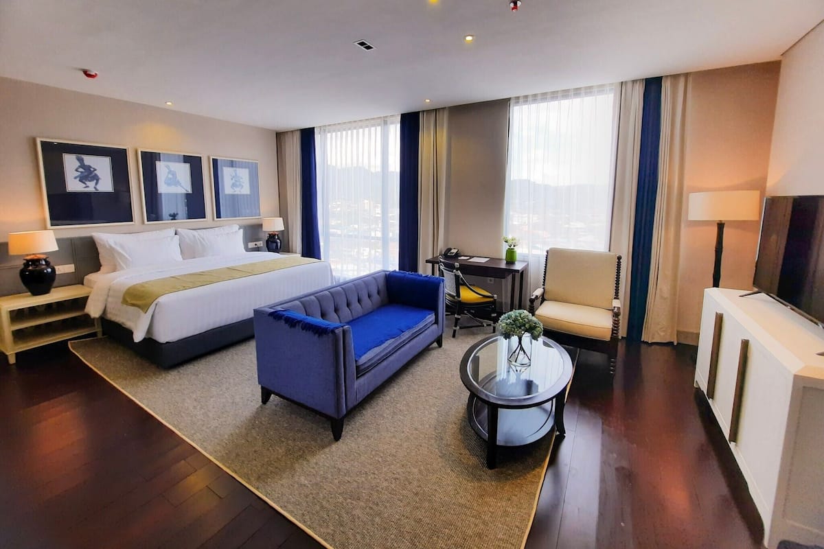 Une chambre d’hôtel avec un lit, un canapé bleu, une petite table avec un vase, une chaise et un téléviseur.