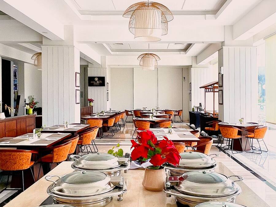 A spacious hotel dining area with multiple tables and chairs, a central decorative vase with red flowers, and a modern ceiling design.