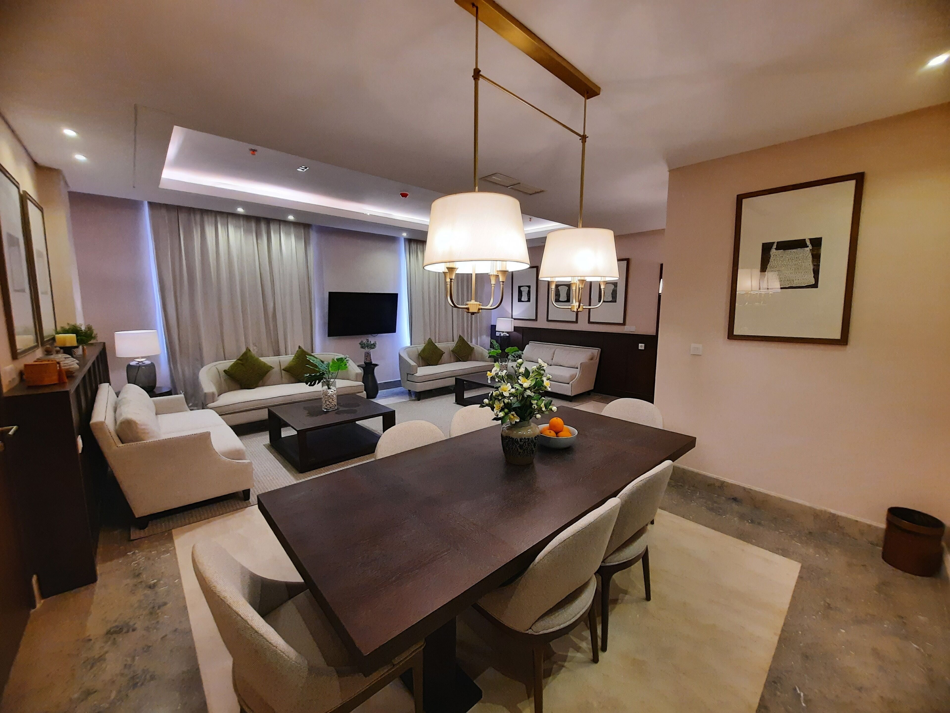 A modern living room with a dining area, featuring a large wooden table, comfortable sofas, and a flat-screen TV.
