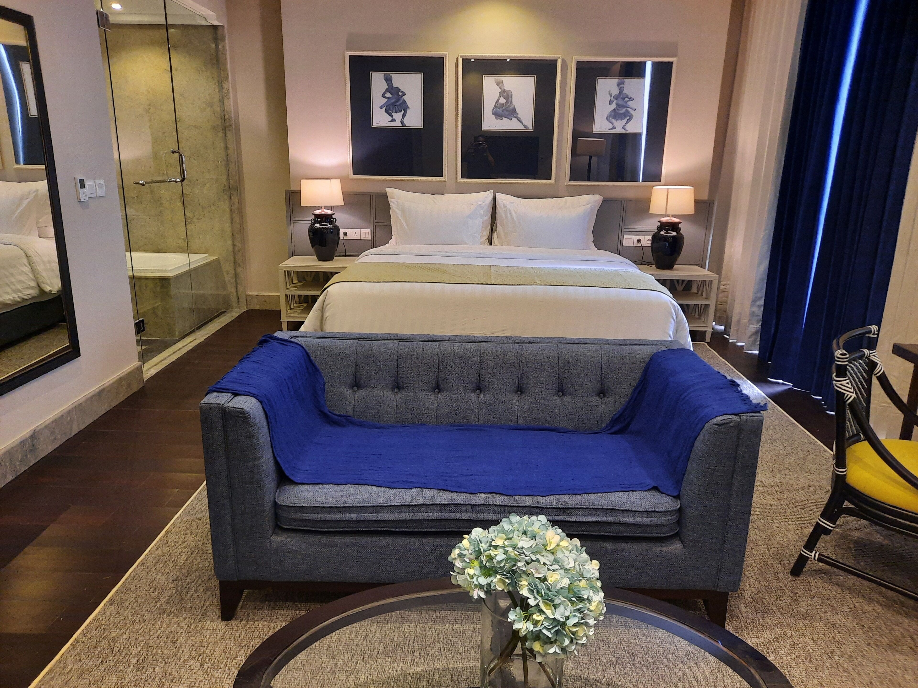 A hotel room with a large bed, two bedside tables, a sofa, a chair, a glass coffee table with flowers, and artwork on the walls.