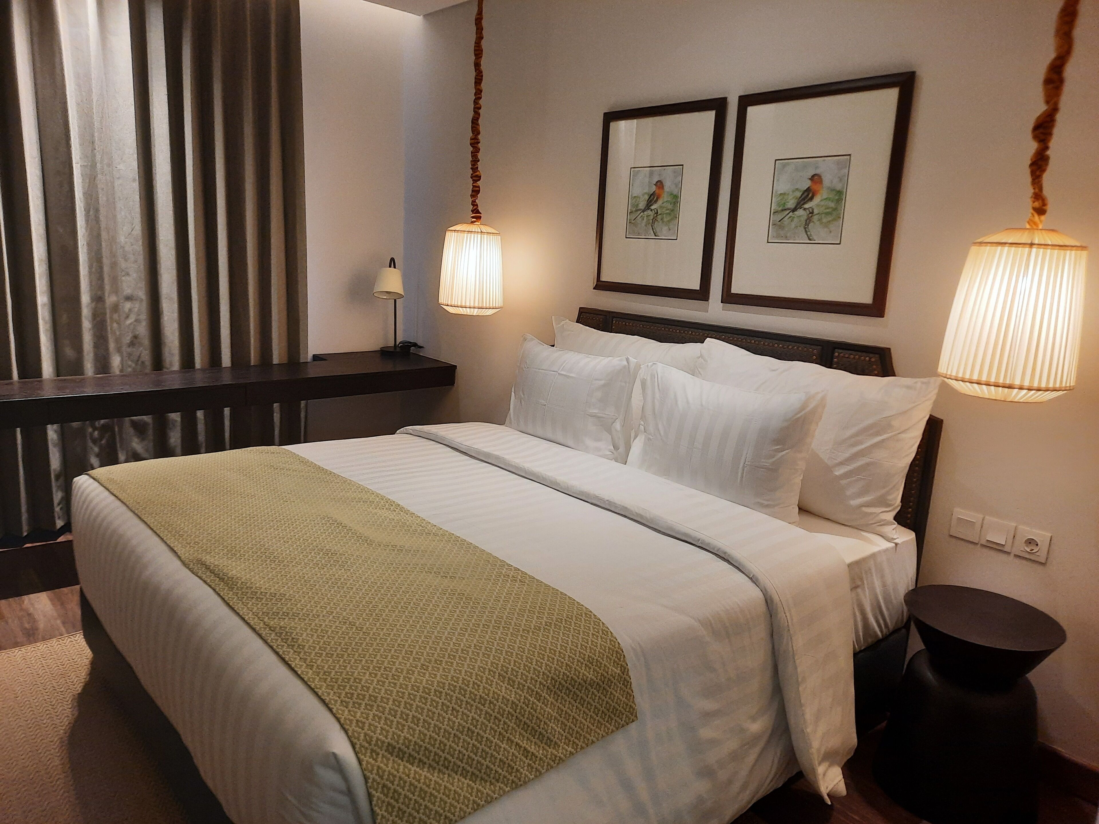Una cama bien hecha con sábanas blancas y un plaid verde, una mesita de noche con lámpara, y dos cuadros en la pared.