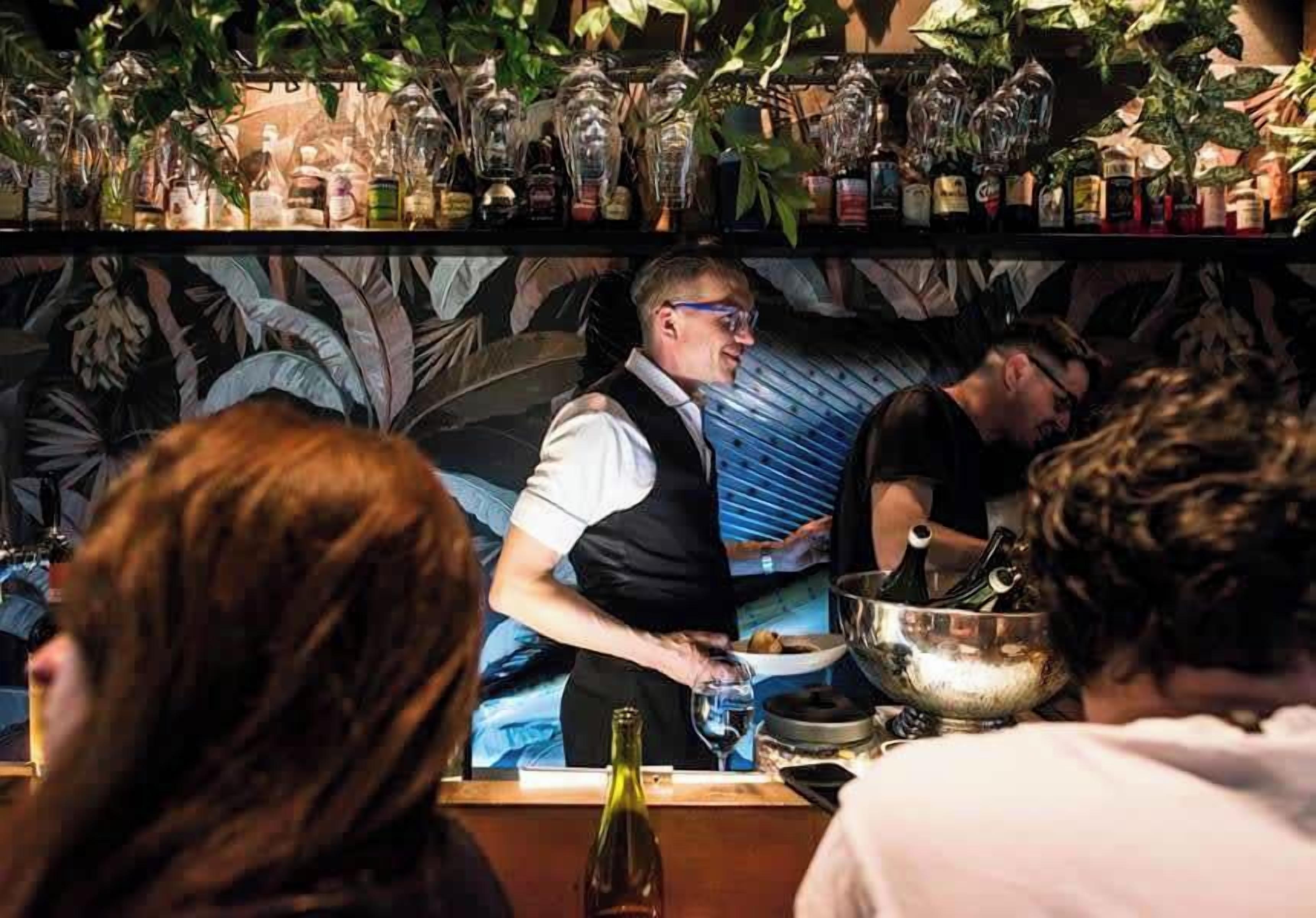 A bar area with a bartender serving drinks, shelves with bottles, and a decorative wall.