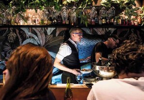 Zona de bar con un cantinero sirviendo bebidas, estantes con botellas y una pared decorativa.
