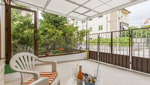 A balcony with a white chair, a small table with a bottle and glasses, and a view of a green fence and buildings. - Comfortable Modern Studio With Shaded Patio (Split)